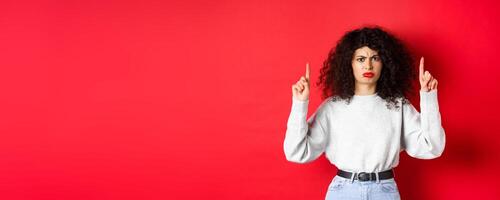 Grumpy young woman with curly hair frowning and grimacing unsatisfied, pointing fingers up at something bad, complaining on company, red background photo