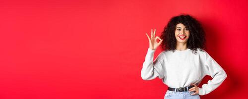 Confident smiling woman in casual clothes, showing OK sign to approve or like product, guarantee good quality, making a compliment, red background photo