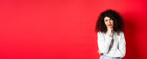 Grumpy young woman with curly hair, looking annoyed or bored at empty space, standing pensive and sad on red background photo
