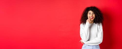 Excited and intrigued woman eager to try something, make tempted look and biting fingernail amused, standing over red background photo