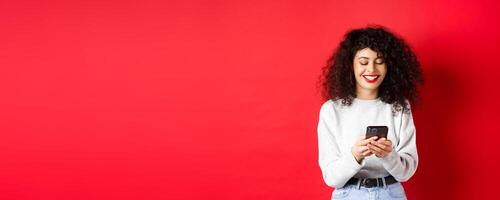 Happy positive girl chatting on phone, reading message and smiling, using social media app, standing on red background photo
