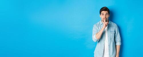 Portrait of excited man asking to keep quiet, showing hush taboo sign and looking nervously at camera, standing against blue background photo