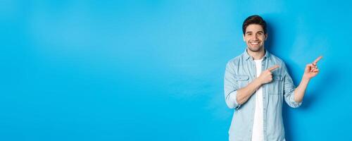 Portrait of attractive adult man smiling, pointing fingers right at logo or banner, showing advertisement against blue background photo
