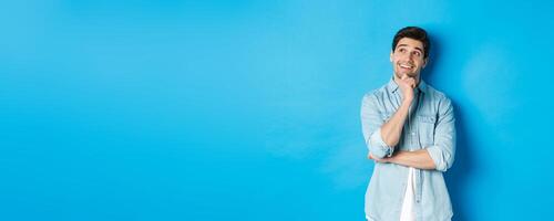 Portrait of thoughtful handsome man with beard, standing in casual outfit, looking at upper left corner and smiling, imaging or dreaming about something, standing over blue background photo
