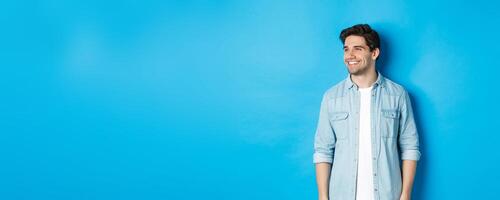 Handsome smiling adult man in casual outfit, smiling and looking left at promo offer, standing against blue background photo