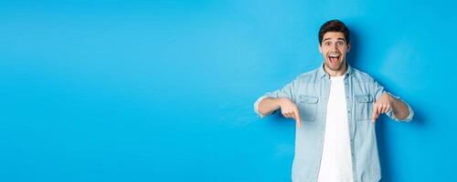 Image of excited handsome man pointing fingers down, making an announcement, standing against blue background photo