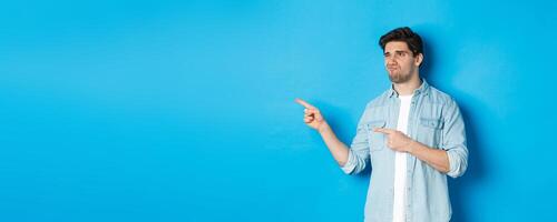 Doubtful adult man pointing fingers left at promotion and looking unsure, grimacing disappointed, standing against blue background photo