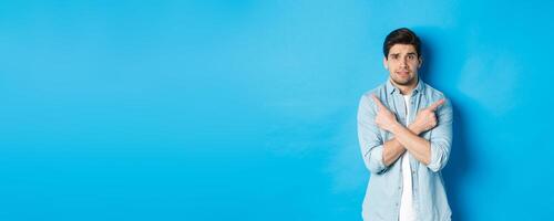 Portrait of nervous guy pointing fingers sideways, looking indecisive and asking for help with choice, showing left and right promo offers, standing against blue background photo
