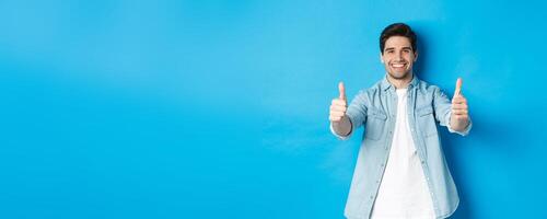 Smiling confident man showing thumbs up, guarantee quality, approving something good, standing against blue background photo