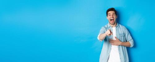 Adult man in casual outfit laughing out loud and pointing at you, looking at something funny, chuckling while standing against blue background photo