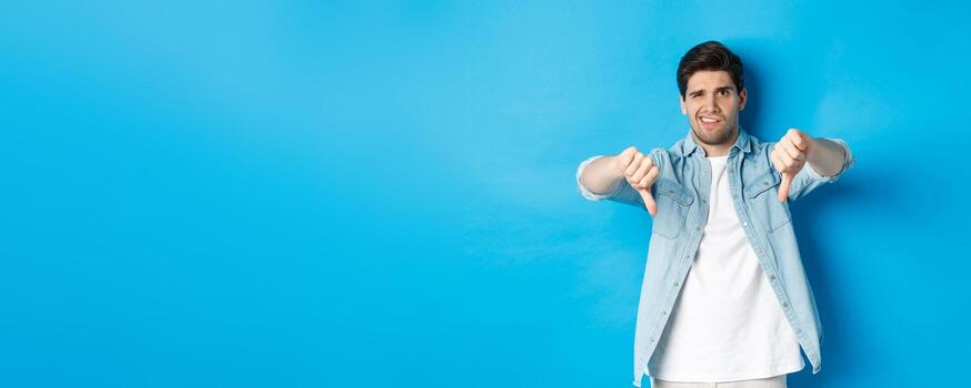 decepcionado hombre demostración pulgares abajo y haciendo muecas a alguna cosa horrible, disgusto producto, juzgando malo anuncio publicitario, en pie en contra azul antecedentes foto