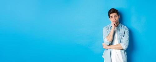 Unamused and bored man looking without interest at camera, standing against bllue background photo