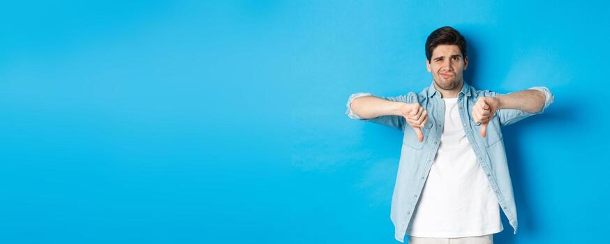 escéptico y decepcionado 25 años antiguo hombre en casual atuendo disgusto alguna cosa malo, demostración pulgares abajo y haciendo muecas disgustado, en pie en contra azul antecedentes foto