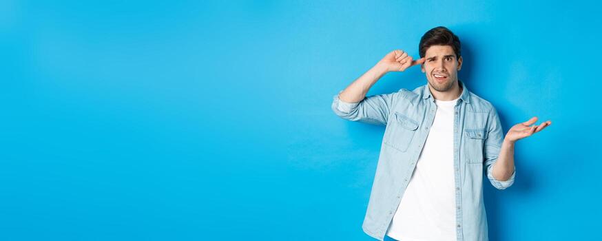 confuso hombre regaño persona para siendo estúpido, señalando dedo a cabeza y encogiéndose de hombros, mirando perplejo, desaprobar comportamiento, en pie en contra azul antecedentes foto