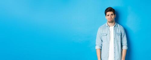 Man looking with doubt at camera, frowning and sulking, feeling uneasy about something, standing against blue background photo