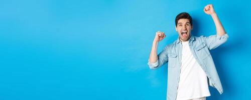 Cheerful guy making fist pumps and rooting for someone, shouting for joy, triumphing over win, standing against blue background photo