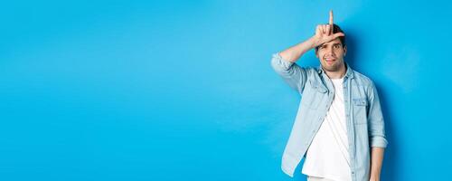 Rude guy mocking person for losing, showing loser sign on forehead and looking with dismay, standing against blue background photo