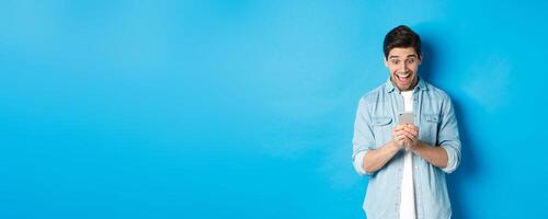 Image of excited man smiling while looking at mobile phone, shopping online on smartphone, standing against blue background photo