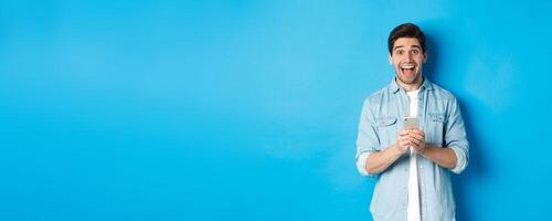 Surprised and happy man winning something online, holding smartphone and rejoicing, standing against blue background photo