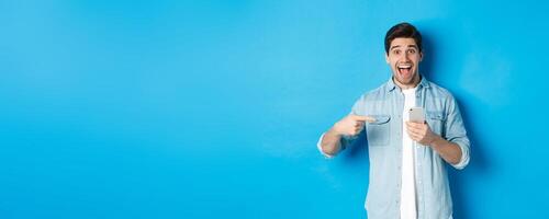 Concept of online shopping, applications and technology. Happy smiling man pointing finger at phone, standing amazed against blue background photo