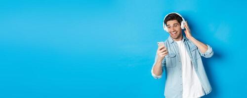 Happy man listening to music in headphones and reading message on smartphone, smiling excited, standing against blue background photo