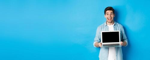 Cheerful smiling man making presentation, showing laptop screen and looking happy, standing over blue background photo