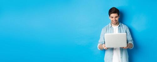 Handsome man working on laptop, smiling and looking at screen satisfied, standing against blue background photo