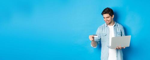 Young man order online, holding credit card and laptop, shopping in internet, standing against blue background photo