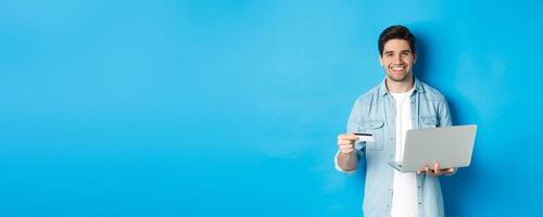 Handsome man smiling, shopping online and paying for product, holding credit card with laptop, standing over blue background photo