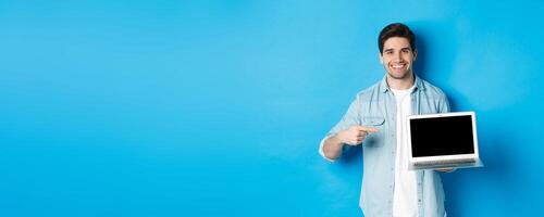 Handsome young man pointing finger at screen of computer, smiling pleased, showing promo in internet or website, standing over blue background photo