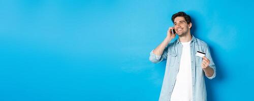 Smiling man call bank support, talking on mobile phone and holding credit card, standing against blue background photo