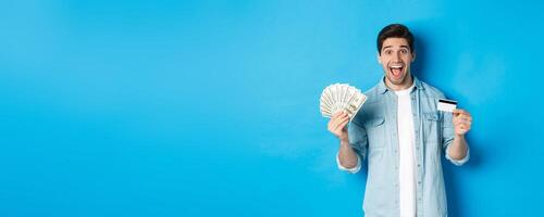 Happy attractive man looking amazed, showing cash and credit card, concept of banks, credit and finance. Blue studio background photo