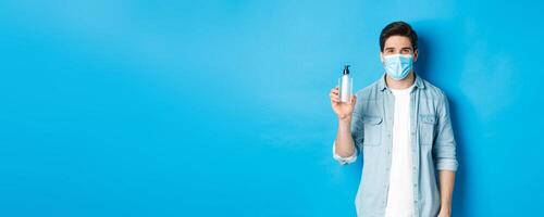 Concept of covid-19, pandemic and social distancing. Young man in medical mask showing hand sanitizer, standing against blue background photo