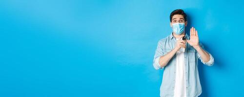 Concept of covid-19, pandemic and social distancing. Cheerful guy in medical mask showing how to disinfect hands with sanitizer, using antiseptic, preventing virus spread, blue background photo