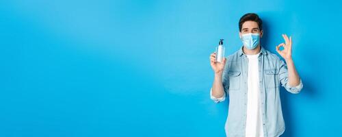 Concept of covid-19, pandemic and social distancing. Satisfied young man in medical mask recommending hand sanitizer, showing ok sign and antiseptic, standing against blue background photo