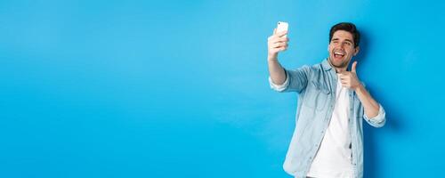 Handsome modern guy taking selfie on smartphone and pointing finger gun at mobile camera, winking cheeky, standing against blue background photo