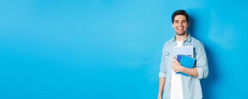 Smiling man studying, holding notebooks and looking happy, standing over blue background photo