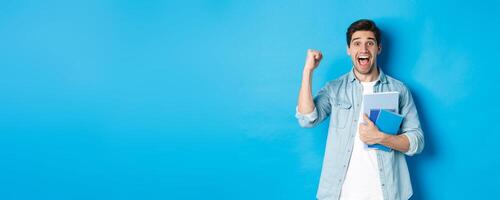 Cheerful guy holding notebooks and celebrating, making fist pump and shouting yes with excitement, standing over blue background photo