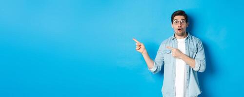 Surprised man in glasses pointing fingers left at copy space, asking question about product, looking intrigued, standing over blue background photo