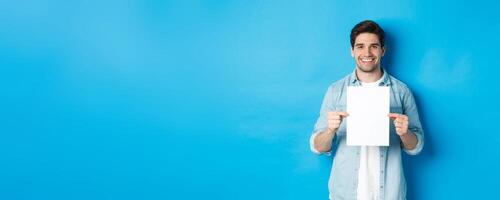 Young smiling guy in casual outfit, holding blank piece of paper with your advertisement, standing over blue background photo