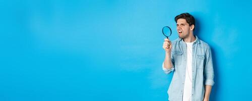 Young thoughtful guy looking through magnifying glass, reading something tiny, standing over blue background photo