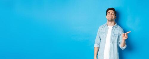 Confident handsome guy smiling sassy, pointing finger right at copy space, showing advertisement, standing over blue background photo