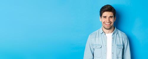 Close-up of young man feeling awkward, smile and cringe from uncomfortable situation, standing over blue background photo