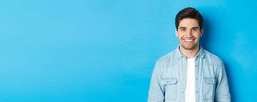 Close-up of young successful man smiling at camera, standing in casual outfit against blue background photo