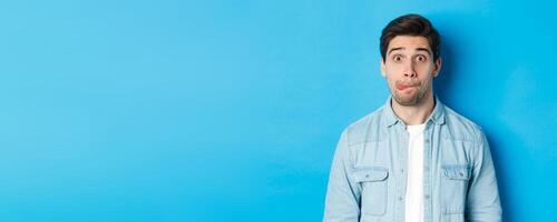 Close-up of young man making funny expressions, showing tongue and looking at camera, standing over blue background photo