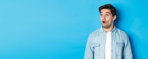 Close-up of surprised handsome man looking impressed left, open mouth fascinated, wearing casual outfit, standing over blue background photo