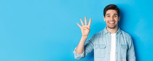 Close-up of handsome man smiling, showing fingers number four, standing over blue background photo