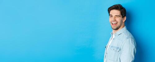 Happy and confident man turn head at camera, winking and smiling, standing over blue background photo
