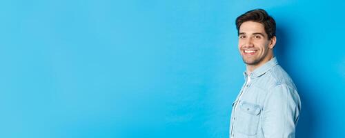 Handsome and confident guy turn head at camera, smiling happy, standing over blue background photo