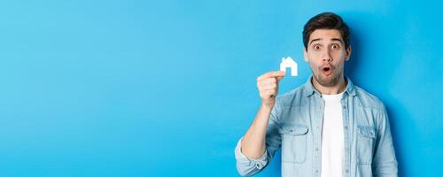 Real estate concept. Surprised guy showing small paper house model, looking amazed, standing against blue background photo
